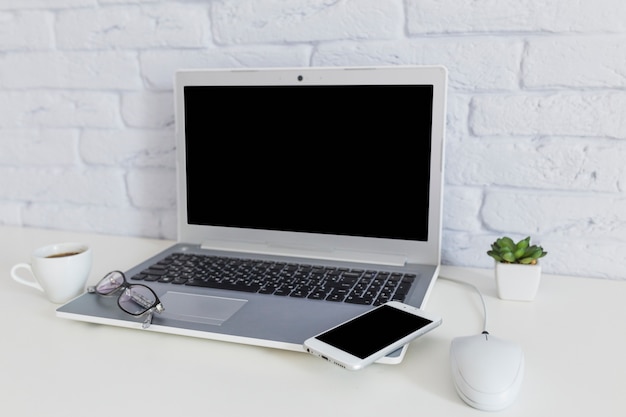Bezpłatne zdjęcie telefon komórkowy i eyeglasses na otwartym laptopie z filiżanką nad białym biurkiem