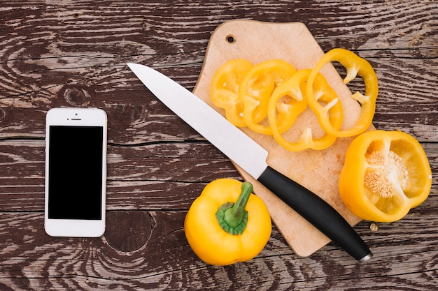 Bezpłatne zdjęcie telefon komórkowy z żółtym dzwonkowym pieprzem i nożem na drewnianym biurku