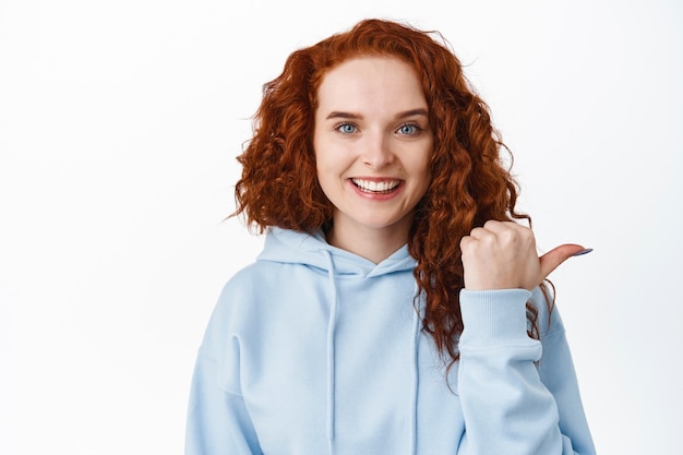 To jest dobra treść. Uśmiechnięta rudowłosa studentka w bluzie z kapturem wskazująca prawą stronę i pokazująca ofertę promocyjną, wyglądająca radośnie na białej ścianie