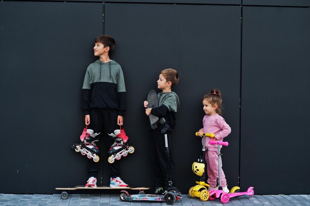 Troje dzieci na świeżym powietrzu przy czarnej nowoczesnej ścianie Sportowa rodzina spędza wolny czas na świeżym powietrzu ze skuterami i łyżwami