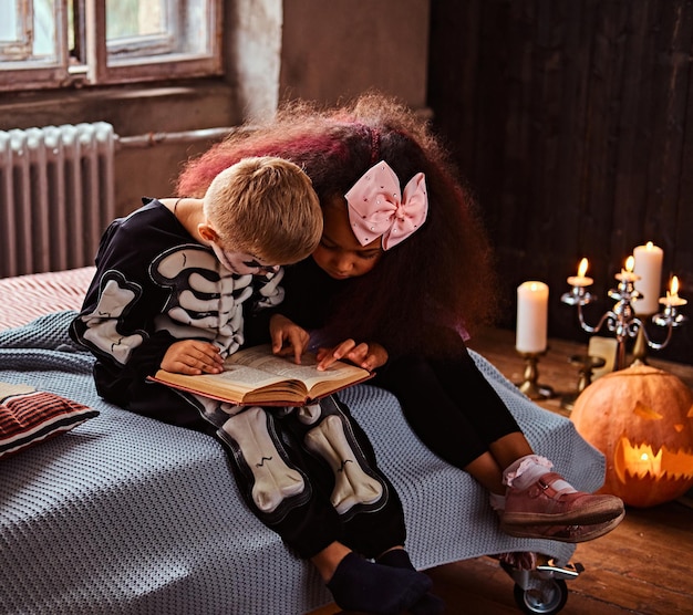 Trójka wielorasowych dzieciaków w przerażających kostiumach czytająca horrory siedząc na łóżku w starym domu. Koncepcja Halloween.