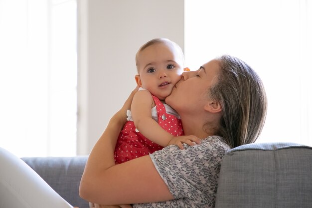 Troskliwa matka siedzi na kanapie i całuje swoją córeczkę z miłością. Urocza dziewczynka a. Długowłosa mama rasy kaukaskiej trzymająca niemowlę obiema rękami. Pojęcie rodziny i macierzyństwa