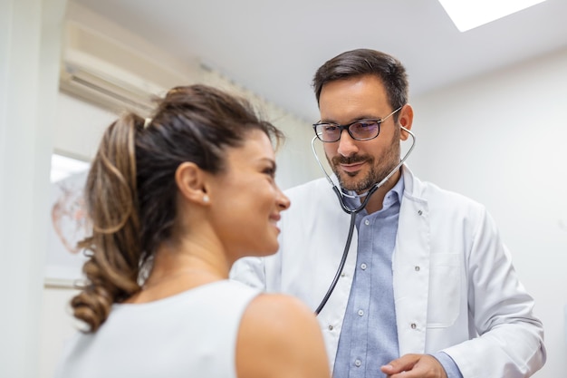 Troskliwy mężczyzna rasy kaukaskiej lekarz używa fonendoskopu bada tętno pacjentki podczas konsultacji w szpitalu Mężczyzna używa stetoskopu lekarz ogólny słuchać bicia serca kobiety w klinice