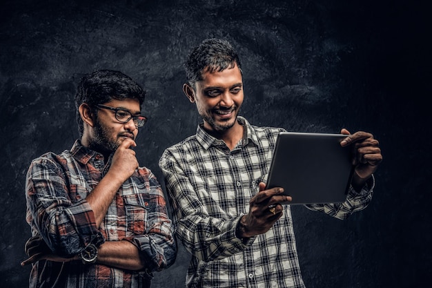 Trzech szczęśliwych indyjskich przyjaciół w zwykłych ubraniach stoi w uścisku i robi selfie na smartfonie. Zdjęcie studyjne na tle ciemnej ściany