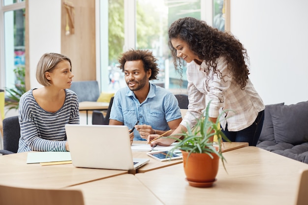Bezpłatne zdjęcie trzej młodzi potencjalni przedsiębiorcy siedzący w bibliotece, omawiający biznesplany i zyski firmy, przeprowadzający badania biznesowe z laptopem, przeglądający informacje na tablecie.