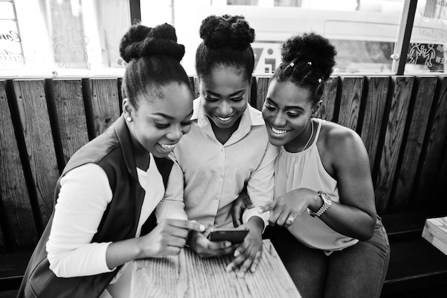 Trzy afroamerykańskie dziewczyny siedzące na stole z kawą i patrzące na telefon komórkowy