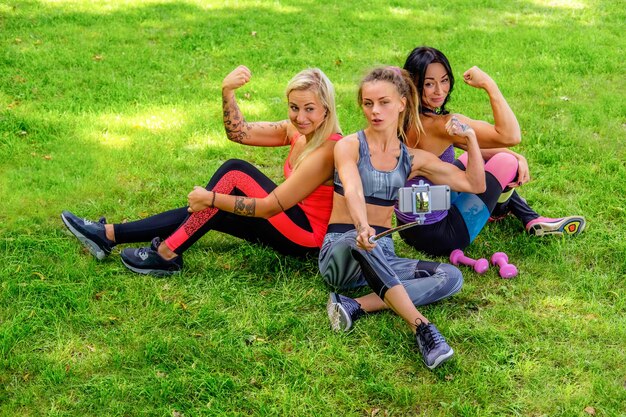 Trzy atrakcyjne, sportowe kobiety siedzące na trawniku i robiące selfie po treningach fitness.