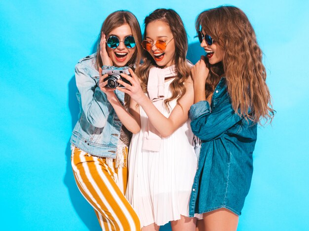 Trzy młode piękne uśmiechnięte dziewczyny w modnych letnich sukienkach i okularach przeciwsłonecznych. Seksowny beztroski kobiet pozować. Robienie zdjęć aparatem retro