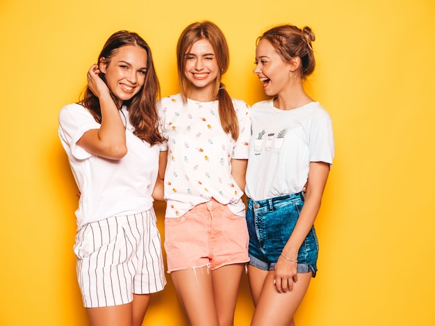 Trzy Młode Piękne Uśmiechnięte Hipster Dziewczyny W Modne Letnie Ubrania. Seksowne Beztroskie Kobiety Pozuje Blisko Kolor żółty ściany. Pozytywne Modele Wariują I Dobrze Się Bawią