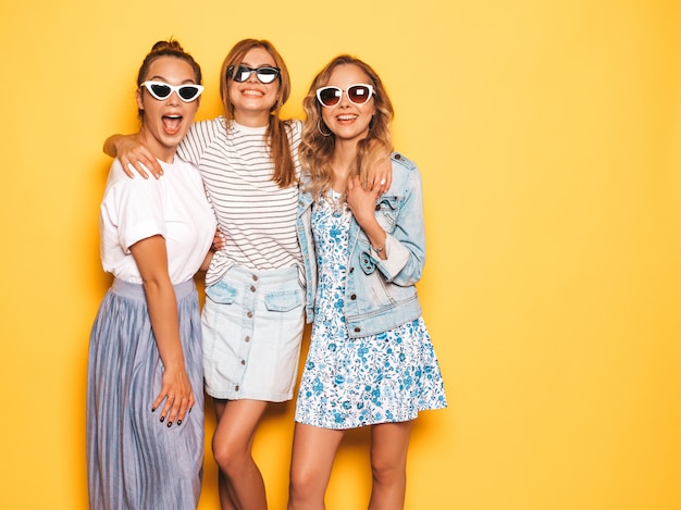 Trzy młode piękne uśmiechnięte hipster dziewczyny w modne letnie ubrania. Seksowne beztroskie kobiety pozuje blisko kolor żółty ściany. Pozytywne modele zabawy w okularach przeciwsłonecznych