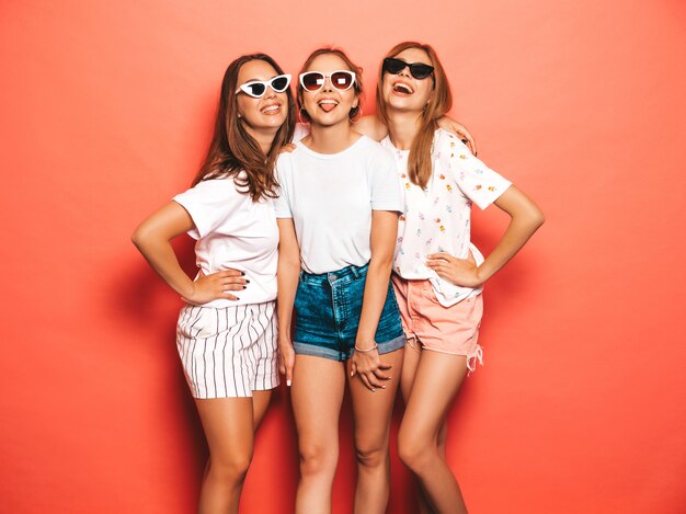 Trzy młode piękne uśmiechnięte hipster dziewczyny w modne letnie ubrania. Seksowne beztroskie kobiety pozuje blisko menchii ściany. Pozytywne modele wariują i dobrze się bawią. W okularach przeciwsłonecznych