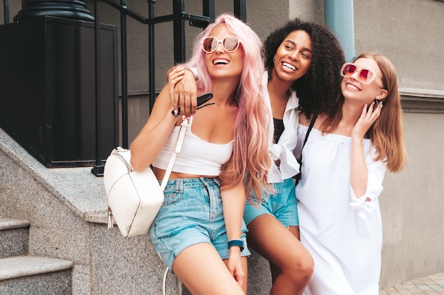 Trzy młode piękne uśmiechnięte kobiety hipster w modnych letnich ubraniachSeksowne beztroskie wielorasowe kobiety pozują na tle ulicyPozytywne modele bawiące się w okularach przeciwsłonecznych Wesoła i szczęśliwa