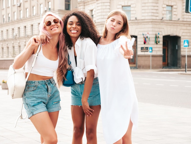 Trzy młode piękne uśmiechnięte kobiety hipster w modnych letnich ubraniachSeksowne beztroskie wielorasowe kobiety pozują na tle ulicyPozytywne modele bawiące się w okularach przeciwsłonecznych Wesoła i szczęśliwa