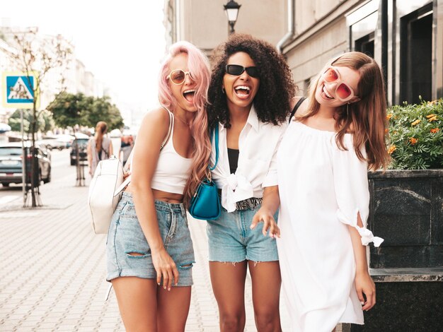 Trzy młode piękne uśmiechnięte kobiety w modnych letnich ubraniachSeksowne beztroskie wielorasowe kobiety pozują na tle ulicyPozytywne modelki bawiące się w okularach przeciwsłonecznych Wesoła i szczęśliwa