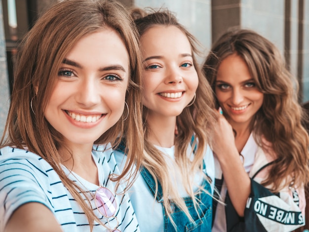 Trzy młode uśmiechnięte kobiety hipster w letnie ubrania. Dziewczyny robienia zdjęć autoportretów na smartfonie. Modele pozowanie na ulicy. Kobieta pokazująca pozytywne emocje na twarzy