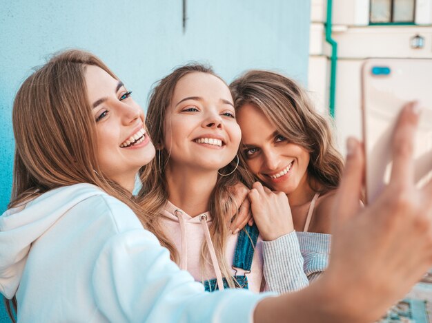 Trzy młode uśmiechnięte kobiety hipster w letnie ubrania. Dziewczyny robienia zdjęć autoportretów na smartfonie. Modele pozowanie na ulicy w pobliżu ściany. Kobieta z pozytywnymi emocjami twarzy