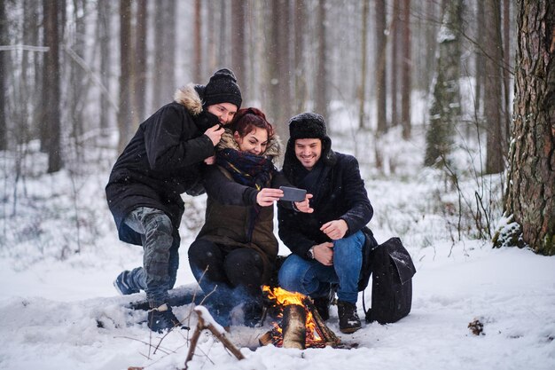 Turyści siedzą przy ognisku, robiąc zdjęcie selfie smartfonem. Wędruj po zimnym, śnieżnym lesie
