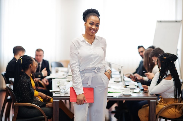 Twarz przystojny african american kobieta biznesu trzymając tablet na tle wielorasowego spotkania zespołu narodów biznesu, siedząc w biurowym stole