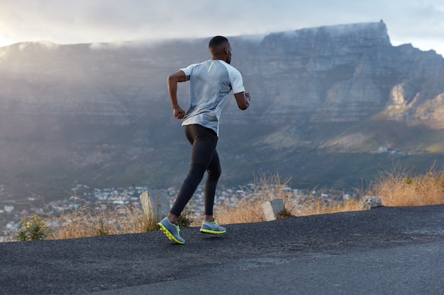 Tylne ujęcie przedstawiające aktywnego ciemnoskórego mężczyznę w akcji, biegnie górską drogą, prowadzi zdrowy tryb życia, ma wytrwałość i motywację do bycia w formie, pozuje w górach, lubi przyrodę