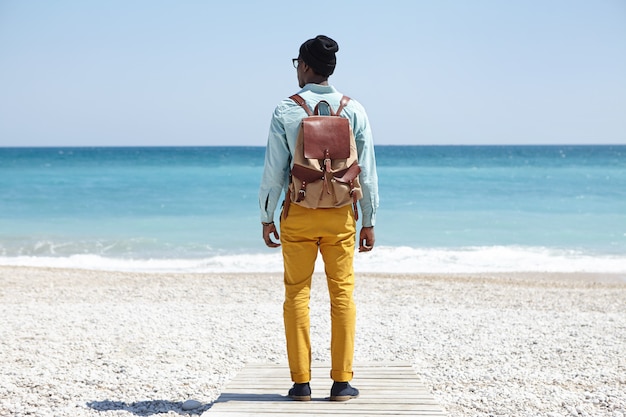 Tylne ujęcie stylowego młodego Afro American backpackera stojącego na promenadzie na kamienistej plaży, z widokiem na rozległy, spokojny ocean z czystą lazurową wodą podczas spokojnego poranka, podziwiając niesamowity widok na morze