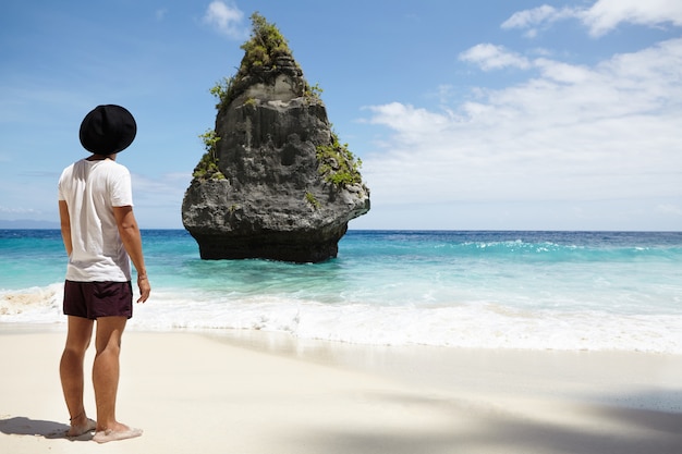 Tylne ujęcie stylowego młodego mężczyzny z bosymi stopami stojącego samotnie na piaszczystej plaży i patrzącego na niesamowitą skalistą wyspę na oceanie, spędzając wakacje w tropikach. Koncepcja ludzi, podróży i przygody