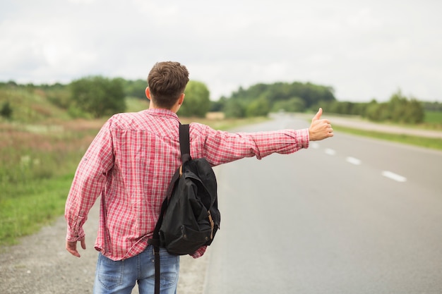 Tylni widok młody człowiek hitchhiking na prostej drodze z jego plecakiem