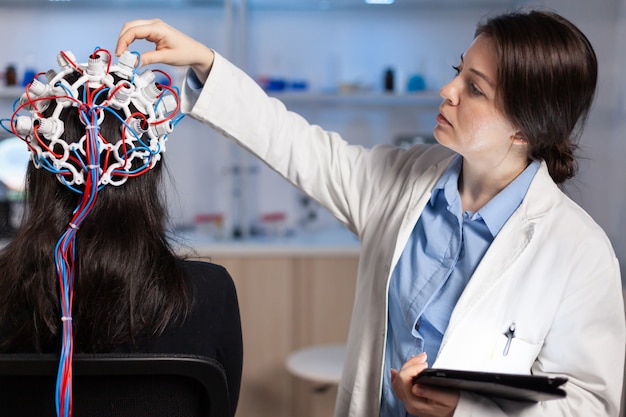 Bezpłatne zdjęcie tylny widok kobiety pacjenta noszenia wydajnego zestawu słuchawkowego eeg siedzącego na krześle w laboratorium badań neurologicznych, podczas gdy badacz medyczny dostosowując go, badając układ nerwowy, wpisując na tablecie.