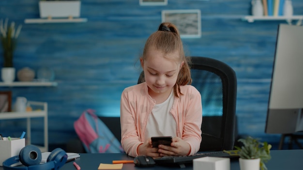 Uczennica patrząc na smartfona do prac domowych i kursów w domu. małe dziecko korzystające z telefonu komórkowego z ekranem dotykowym do zdalnych lekcji online i edukacji przy biurku. uczeń uczący się