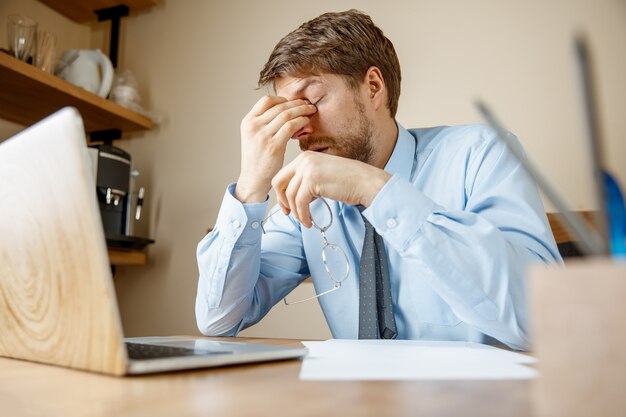 Uczucie mdłości i zmęczenia. Sfrustrowany smutny nieszczęśliwy chory młody człowiek masuje głowę, siedząc w swoim miejscu pracy w biurze.