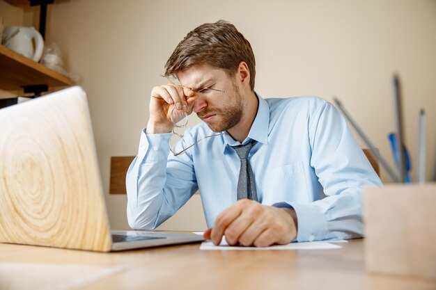 Uczucie mdłości i zmęczenia. Sfrustrowany smutny nieszczęśliwy chory młody człowiek masuje głowę, siedząc w swoim miejscu pracy w biurze.