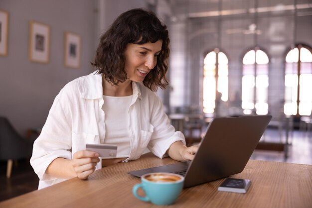 Udana młoda brunetka kaukaska dokonuje płatności online za pomocą karty kredytowej i laptopa siedzącego przy stole