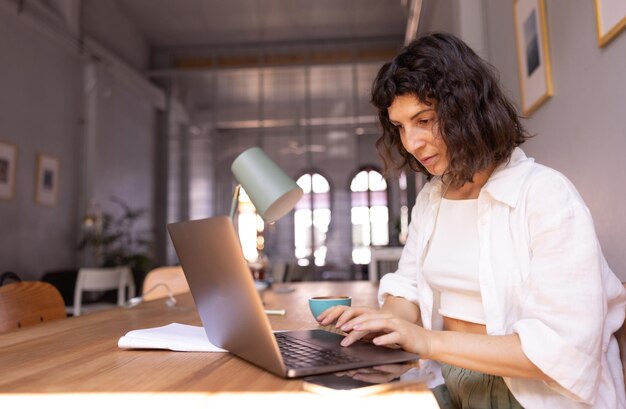 Udana młoda brunetka kaukaska dziewczyna w białej koszuli wpisuje tekst na laptopie, siedząc przy stole w pomieszczeniu