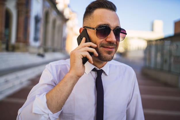 Udany młody biznesmen rozmawia przez telefon w formalnym stroju z okularami przeciwsłonecznymi