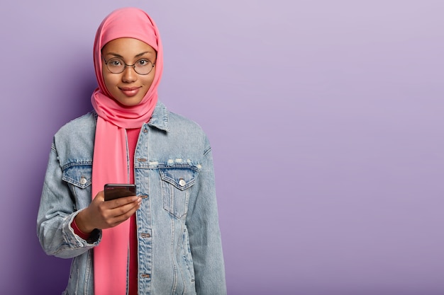 Ujęcie dobrze wyglądającej ciemnoskórej kobiety o atrakcyjnym wyglądzie nosi różowy hidżab i dżinsowy płaszcz, trzyma nowoczesny telefon komórkowy, czeka na ważny telefon, stoi nad fioletową ścianą z pustą przestrzenią