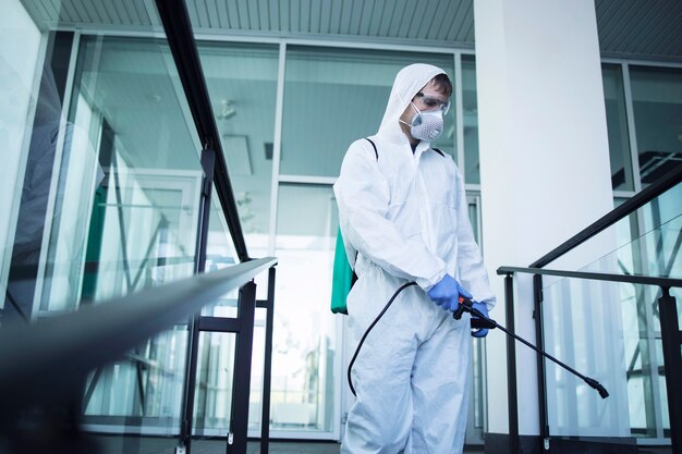 Ujęcie mężczyzny w białym kombinezonie chroniącym przed chemikaliami, wykonującego dezynfekcję miejsc publicznych w celu powstrzymania rozprzestrzeniania wysoce zaraźliwego wirusa koronowego