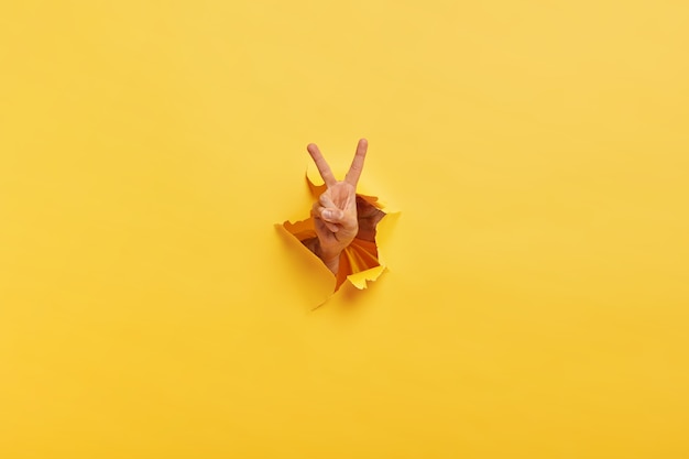 Bezpłatne zdjęcie ujęcie nierozpoznawalnego człowieka pokazuje znak zwycięstwa przez rozdarty otwór w żółtym papierze