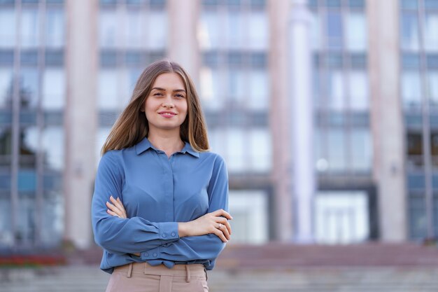 Ujęcie pięknej młodej bizneswoman na sobie niebieską koszulę z szyfonu, stojąc na budynku na ulicy z założonymi rękami.