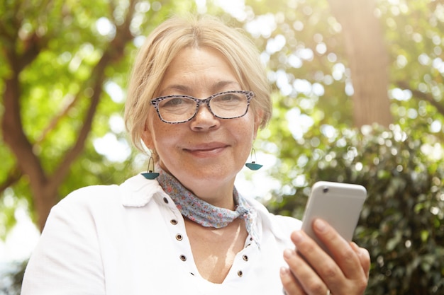 Bezpłatne zdjęcie ujęcie w głowę pięknej kaukaskiej rencistki wyglądającej z radosnym, wesołym wyrazem twarzy podczas korzystania z telefonu komórkowego do komunikowania się online ze znajomymi, czytania wiadomości, wysyłania zdjęć