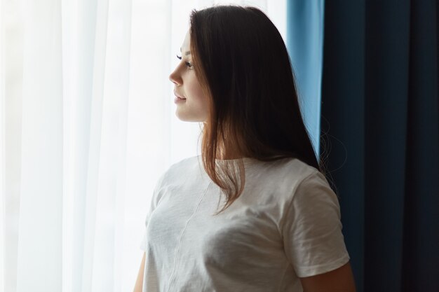 Ujęcie z boku zamyślonej ciemnowłosej kobiety ubranej w białą koszulkę, myśli o czymś stojącym przy oknie