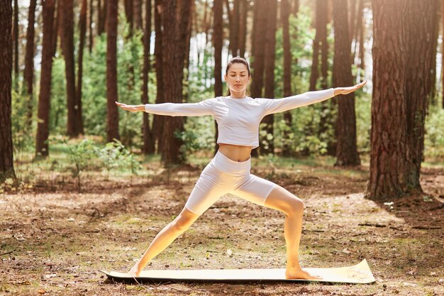 Ujmująca kobieta w białej odzieży sportowej ćwicząca jogę w zielonym parku lub lesie, stojąca w pozycji jogi, trzymająca zamknięte oczy, rozkładająca ręce na bok, trenująca na świeżym powietrzu.