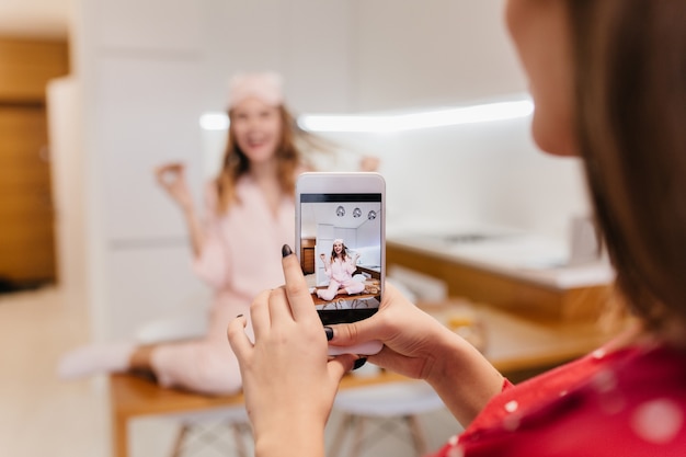 Uradowana biała dziewczyna je pizzę i bawi się włosami. Brunetka dama trzyma smartfon i robi zdjęcie przyjaciela w kuchni z jasnym wnętrzem.