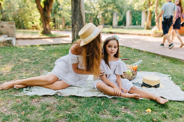 Uradowana ciemnowłosa dziewczyna siedzi na kocu obok matki i dotyka jej nogi. Zewnątrz portret rodziny modnej młodej kobiety i ładnej córki w białej sukni, pozowanie na trawie z ludźmi.