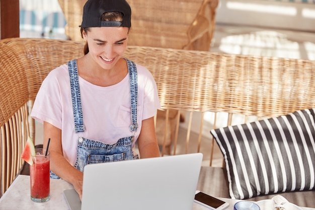 Uradowana hipsterka o wesołym wyrazie twarzy, nosi czapkę i dżinsowy kombinezon, siedzi przed otwartym laptopem, pije świeży letni koktajl, lubi komunikację online i bezpłatny internet