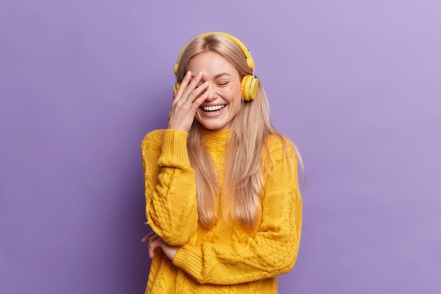 Uradowana młoda Europejka o blond włosach, głośno się śmieje, sprawia, że dłoń słucha muzyki przez słuchawki bezprzewodowe, nosi swobodny żółty sweter