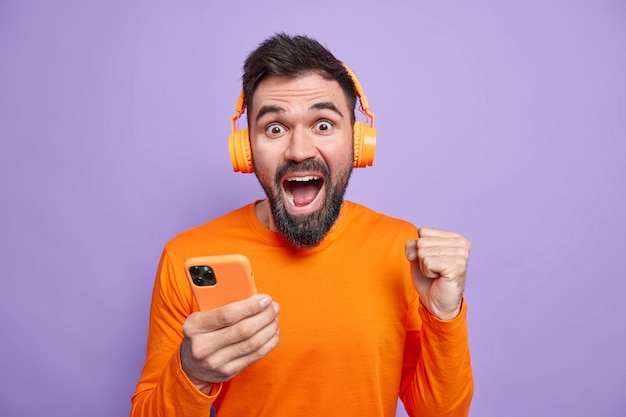 Uradowany nieogolony mężczyzna świętuje wspaniałe wieści zaciska pięści w dłoniach telefon komórkowy słucha muzyki słucha w słuchawkach bezprzewodowych cieszy się dobrym dźwiękiem nosi pomarańczowy sweter