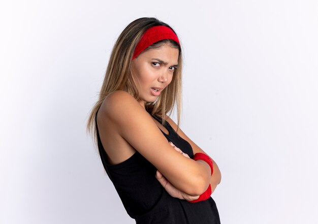 Urażona młoda dziewczyna fitness w czarnej odzieży sportowej i czerwonej opasce na głowę stojącej nad białą ścianą