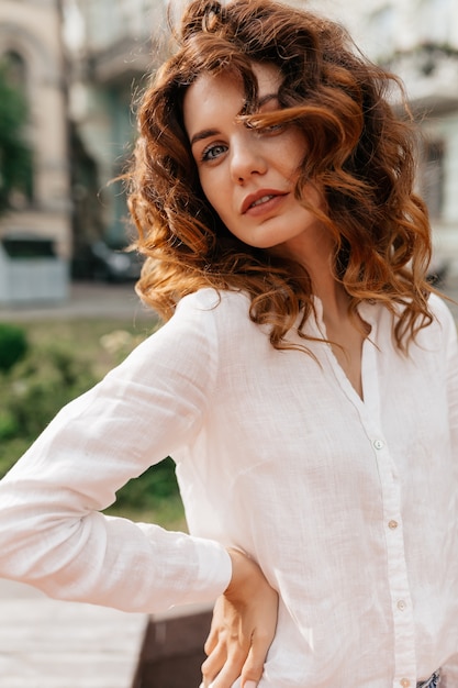 Urocza atrakcyjna kobieta kręcone w białej koszuli pozuje z uśmiechem na zewnątrz podczas sesji zdjęciowej