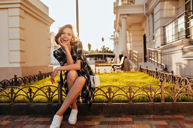 Bezpłatne zdjęcie urocza biała dziewczyna w półbutach gumowych pozuje w słoneczny jesienny dzień zewnątrz zdjęcie stylowej jocund modelki siedzącej na ulicy