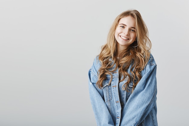 Urocza blond dziewczyna uśmiechnięta szczęśliwa w dżinsowej kurtce, stojąca na szarej ścianie