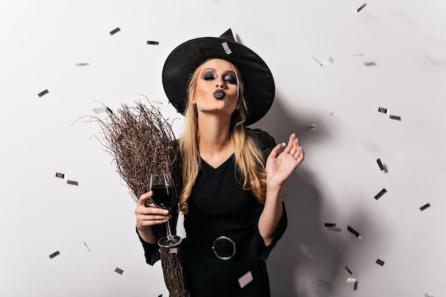 Urocza blond wiedźma delektująca się winem. Wesoła jasnowłosa dama w kostiumie maskarady pozuje na imprezie z okazji halloween.